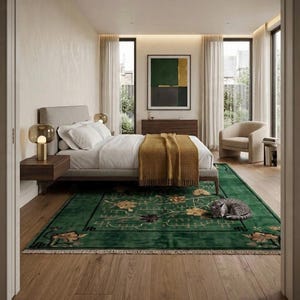Op de afbeelding: Slaapkamerinterieur met een bed met wit beddengoed en een mosterdkleurige deken. Een groen tapijt met bloemmotieven en een slapende grijze kat op de voorgrond. De kamer heeft grote ramen, een stoel en een kunstwerk.