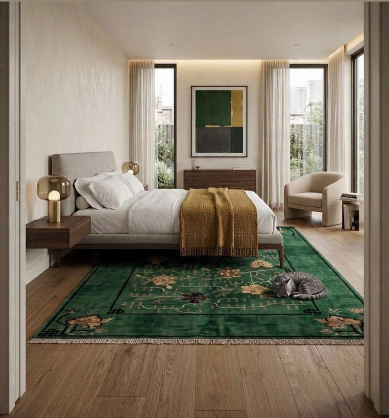 Op de afbeelding: Slaapkamerinterieur met een bed met wit beddengoed en een mosterdkleurige deken. Een groen tapijt met bloemmotieven en een slapende grijze kat op de voorgrond. De kamer heeft grote ramen, een stoel en een kunstwerk.