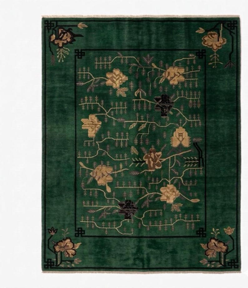 Op de afbeelding: Een rechthoekig groen tapijt met een bloemenontwerp. Het tapijt heeft een donkergroene rand en hoekaccenten met een zwart geometrisch patroon. Het centrale ontwerp omvat gouden en zwarte bloemen- en takmotieven.
