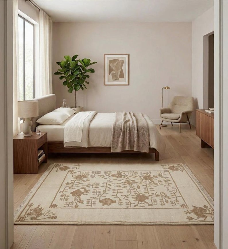 May include: A bedroom with a bed featuring cream bedding and a brown wooden frame. A beige rug with a floral design is on the wooden floor. A side table with a lamp and a potted plant complete the room's decor, creating a calm atmosphere.