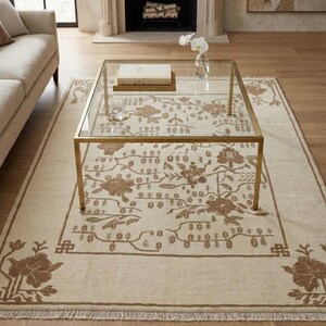 May include: A beige rectangular rug featuring a brown floral design. A gold-framed glass coffee table is placed on the rug, with a book and a glass on top. The rug has a fringed edge and a floral border.