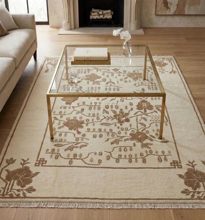 May include: A beige rectangular rug featuring a brown floral design. A gold-framed glass coffee table is placed on the rug, with a book and a glass on top. The rug has a fringed edge and a floral border.