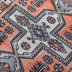 Puede incluir: Primer plano de una alfombra estampada con un diseño geométrico. La alfombra presenta una forma central en forma de cruz en blanco roto, rodeada de patrones geométricos naranjas y grises. El diseño de la alfombra incluye formas y bordes repetidos.