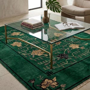 Op de afbeelding: Een moderne woonkamer met een salontafel met glazen blad op een groen tapijt met bloemmotieven. De tafel heeft goudkleurige poten en draagt boeken, een vaas en een plant. Op de achtergrond is een fauteuil in neutrale tinten te zien.