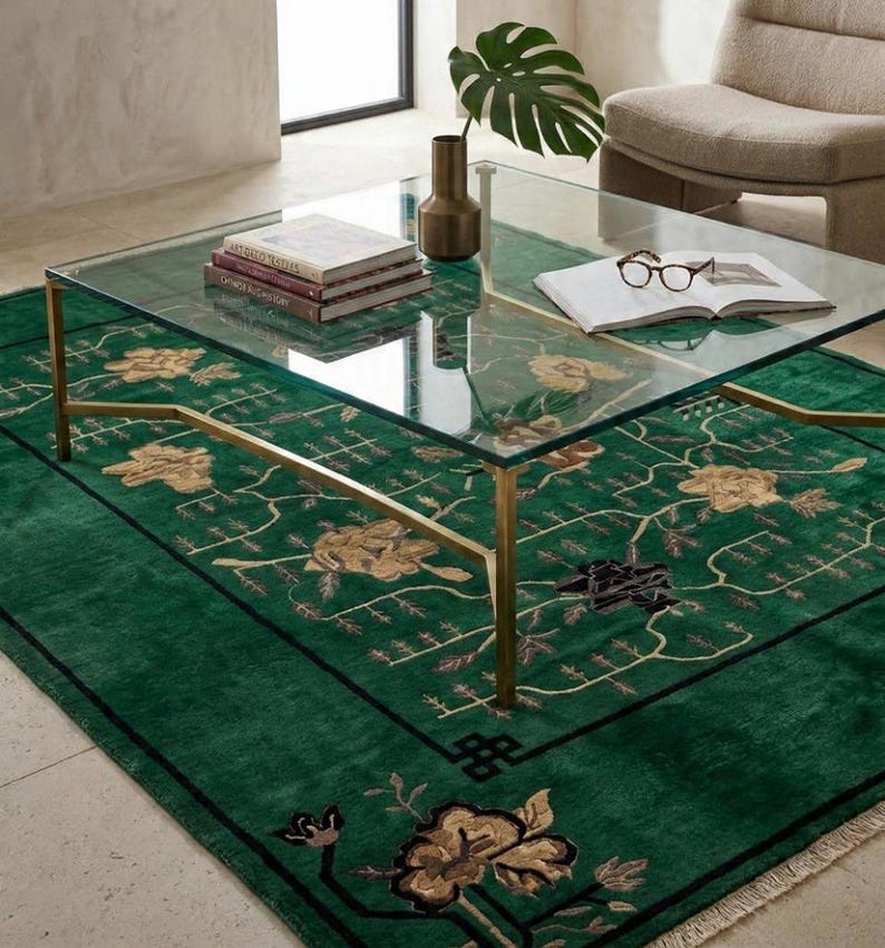 Op de afbeelding: Een moderne woonkamer met een salontafel met glazen blad op een groen tapijt met bloemmotieven. De tafel heeft goudkleurige poten en draagt boeken, een vaas en een plant. Op de achtergrond is een fauteuil in neutrale tinten te zien.
