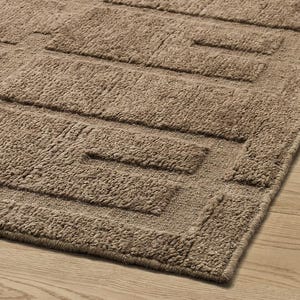 Peut inclure: Gros plan sur un tapis de bain rectangulaire marron avec un motif géométrique. Le tapis a une surface texturée et moelleuse avec des formes rectangulaires en relief. Le tapis est fait d'un matériau doux et est posé sur un sol en bois clair.