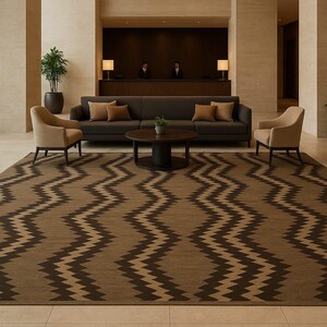 Puede incluir: Un vestíbulo de hotel con una gran alfombra marrón y beige con estampado de zigzag. La habitación incluye un sofá gris oscuro con cojines marrones, dos sillones beige y una mesa de centro redonda. Las plantas y las lámparas completan la decoración.