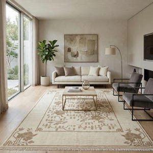 May include: A modern living room with a cream-colored sofa, throw pillows, and a patterned rug. The rug features floral designs. Two grey armchairs with black frames and a coffee table are also present. The room has a neutral colour scheme and a large window.