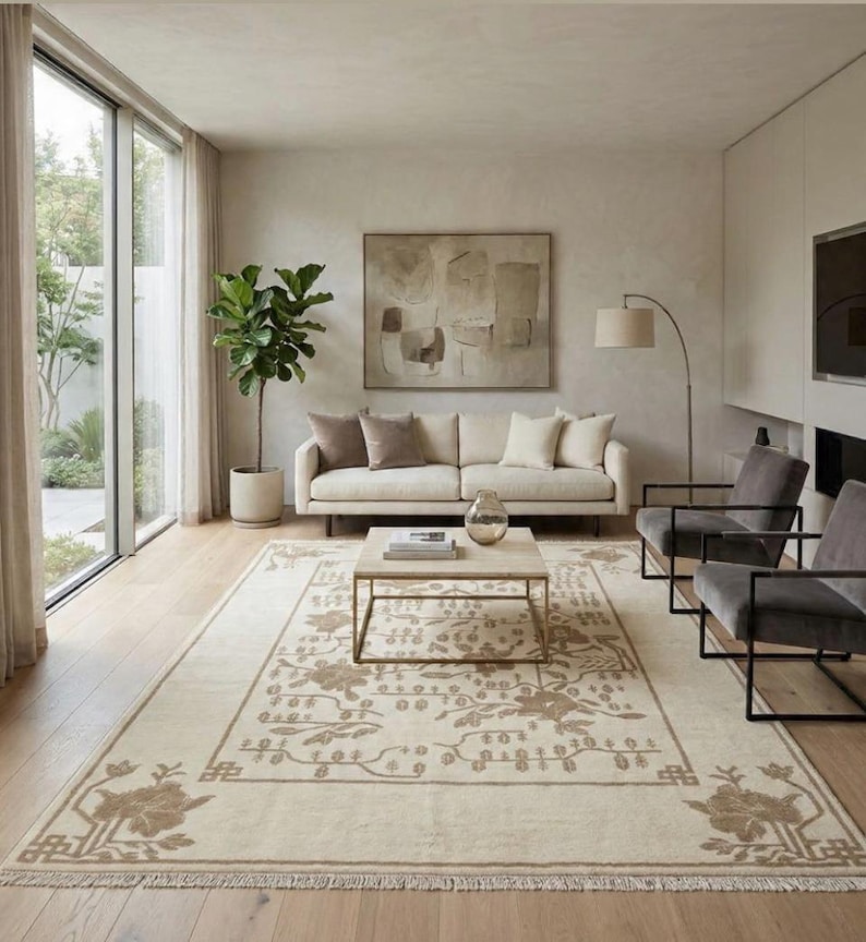 May include: A modern living room with a cream-colored sofa, throw pillows, and a patterned rug. The rug features floral designs. Two grey armchairs with black frames and a coffee table are also present. The room has a neutral colour scheme and a large window.