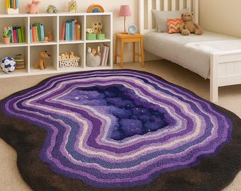 Puede incluir: Una alfombra grande con forma de geoda con un borde exterior marrón oscuro y capas concéntricas de púrpura, lavanda y blanco. El centro es azul oscuro con motas blancas, que se asemejan a una noche estrellada. Esta alfombra es un revestimiento decorativo para el suelo.