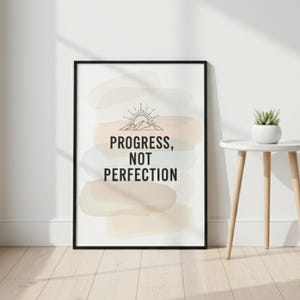May include: A framed art print with the words "PROGRESS, NOT PERFECTION" in black. The artwork features a sunrise design above the text and pastel watercolor-like shapes in the background. A small white side table with a potted plant is next to the artwork.