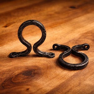 Puede incluir: Dos soportes decorativos de metal oscuro, forjados a mano, con un acabado texturizado. Uno está en posición vertical y el otro está tumbado. Los soportes están sobre una superficie de madera. Tienen un diseño curvo único.