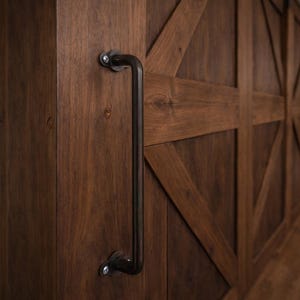 Puede incluir: Armario de madera marrón oscuro con un tirador de metal negro. Las puertas del armario tienen un diseño de travesaño cruzado. El tirador está fijado con tornillos plateados. La veta de la madera es visible.