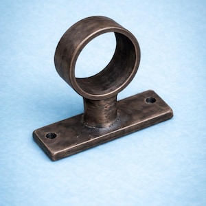 Puede incluir: Un soporte de barra de cortina de color bronce. El soporte tiene un anillo circular unido a una base rectangular con dos orificios para tornillos. El metal tiene un acabado texturizado, sobre un fondo azul claro.