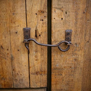 Puede incluir: Primer plano de una puerta de madera rústica con un pestillo de gancho y ojo de metal oscuro. La puerta está hecha de tablones de madera desgastados con vetas y nudos visibles. La ferretería metálica está sujeta con tornillos.