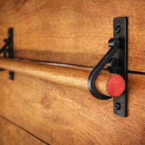 May include: A wooden towel rail with a natural finish and black metal hardware. The rail features a wooden dowel held by decorative black brackets. A small red wooden accent is visible on the right side. The rail is mounted on a wooden surface.