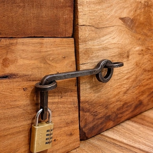 Puede incluir: Un pestillo de metal oscuro hecho a mano con un candado de combinación que asegura dos tablones de madera. El candado es de color dorado y la madera tiene una veta natural rica. El pestillo es una pieza decorativa y funcional.