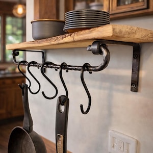Könnte beinhalten: Ein rustikales Küchenregal mit natürlicher Kante, das einen Stapel grauer Teller und eine kleine Schüssel trägt. Darunter hängt eine schwarze Metallstange mit S-Haken, an der zwei schwarze Bratpfannen befestigt sind. Das Design kombiniert Holz und Metall.