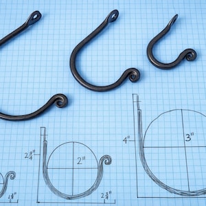 Könnte beinhalten: Drei dunkle Metallhaken mit einem geschwungenen Design, dargestellt auf blauem Rasterhintergrund. Die Haken sind unterschiedlich groß, mit einem Bauplan, der die Abmessungen in Zentimetern zeigt. Das Design beinhaltet eine Schlaufe zum Aufhängen.