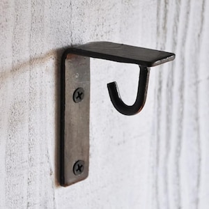 Op de afbeelding: Een zwarte metalen wandhaak met een klein plankje. De haak is met twee schroeven aan een rechthoekige plaat bevestigd. Het plankje steekt horizontaal uit en de haak is naar beneden gebogen. De afwerking is donker en rustiek.