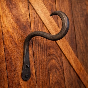 May include: A rustic, hand-forged iron hook with a curved design. The hook is mounted on a wooden door with a warm, brown finish. The hook's dark color contrasts with the wood's lighter tones, creating a visually appealing accent.