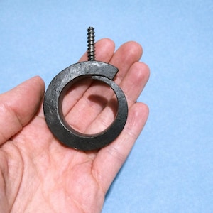 May include: A dark gray, forged metal curtain ring with a screw attached. The ring is circular with a small gap and a textured surface. The screw is silver and extends upwards from the ring.