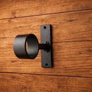 Puede incluir: Un soporte de barra de cortina de metal negro con un anillo circular y una placa de montaje rectangular. Montado sobre una superficie de madera con vetas visibles.