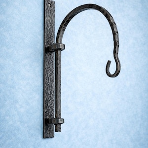 Puede incluir: Un gancho de pared de hierro forjado de color marrón oscuro con un acabado texturizado. El gancho tiene un brazo curvado y un extremo rizado, diseñado para colgar objetos. La placa de montaje es rectangular y tiene un agujero.