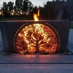 May include: A rectangular, dark metal fire pit with a tree design cut out of the front. Flames are visible inside the pit, and the fire pit is sitting on a bed of small rocks. The background shows trees and a dusky sky.