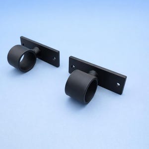 Może przedstawiać: Dwa matowe, czarne wsporniki do karniszy. Każdy wspornik ma prostokątną podstawę z dwoma otworami na śruby i okrągły pierścień. Wsporniki są umieszczone na jasnoniebieskim tle.