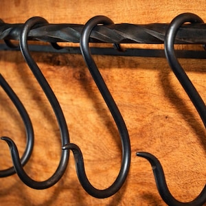 以下が含まれることがあります： S字型のフックがいくつか付いた黒い金属製の鍋ラックのクローズアップ。ラックはねじれた金属棒で、木製の表面に取り付けられています。フックは等間隔に配置されています。