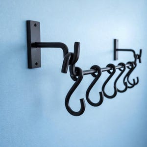 Puede incluir: Estante de metal negro para colgar en la pared con seis ganchos en forma de S. El estante tiene una barra horizontal con los ganchos y dos soportes de montaje. El diseño es simple y funcional, adecuado para la cocina o el almacenamiento.