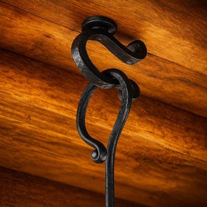 Puede incluir: Un gancho de hierro forjado negro, hecho a mano, con un diseño decorativo en espiral. El gancho está montado sobre una superficie de madera con una veta natural y cálida. El diseño incluye un bucle y un extremo curvado.