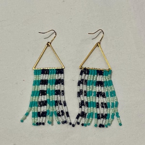 Puede incluir: Un par de pendientes de cuentas con una parte superior triangular dorada y flecos. Los flecos están hechos de cuentas turquesas, negras, blancas y verde claro. Los pendientes están diseñados para colgar.