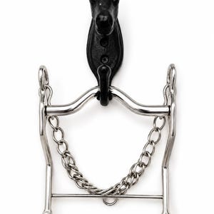 May include: A silver-coloured metal horse bit with a curb chain, suspended from a black cast iron wall hook shaped like a horse head. The bit features two side shanks with rings at the bottom and a curved mouthpiece, all hanging vertically.