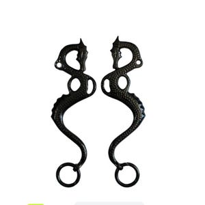 Puede incluir: Dos bocados de caballo negros con un diseño decorativo. Cada bocado tiene una forma curva con una serie de agujeros y un anillo en la parte inferior. Los bocados son de metal y están diseñados para actividades ecuestres.
