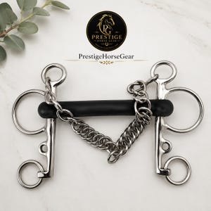 May include: A silver-coloured metal horse bit featuring a straight black rubber mouthpiece and a stainless steel curb chain. The bit includes two side shanks with multiple rings and the logo Prestige Horse Gear printed above the item.
