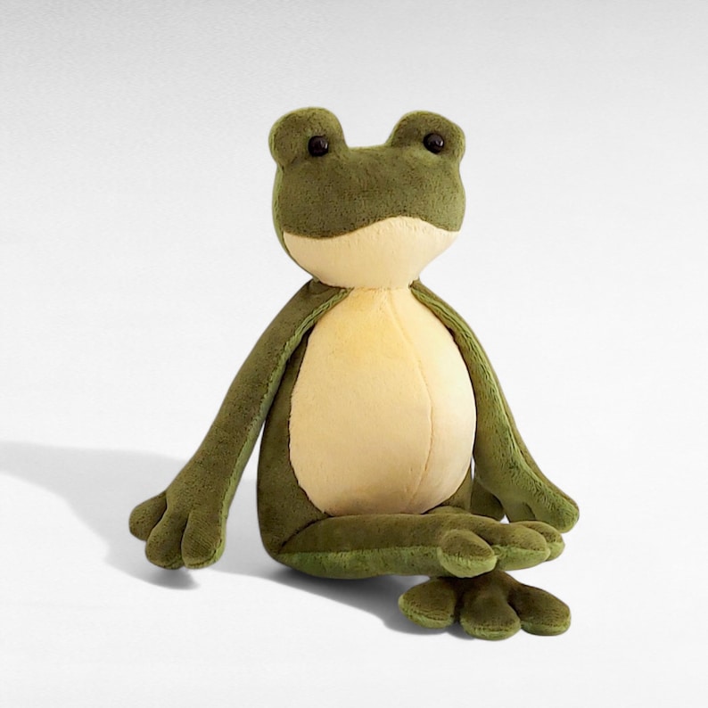 May include: A plush frog toy in shades of green and cream. The stuffed animal has large, round eyes and outstretched arms and legs. The frog is sitting on a white surface.