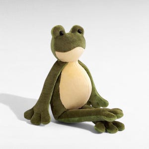 May include: A plush frog toy in shades of green and cream. The frog has a rounded head with black eyes, long arms, and webbed feet. The soft toy is designed to sit upright, with its arms and legs extended.