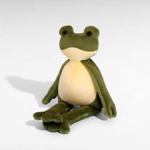 May include: A plush frog toy with a light green body, a cream-colored belly, and dark green arms and legs. The frog has large, round black eyes and is sitting on a white surface. The toy is soft and cuddly.
