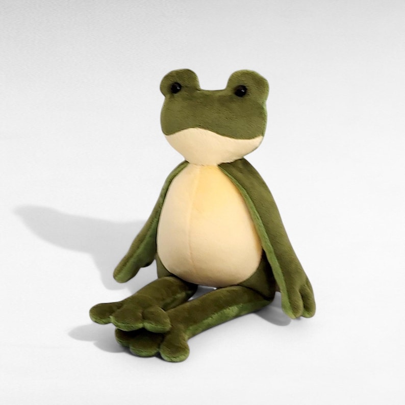 May include: A plush frog toy with a light green body, a cream-colored belly, and dark green arms and legs. The frog has large, round black eyes and is sitting on a white surface. The toy is soft and cuddly.