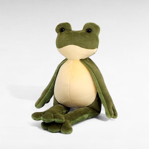 May include: A plush frog toy in shades of green and cream. The frog has a textured, soft body, with a green head, arms, and legs. The frog's belly is cream-colored, and it has black eyes. The frog is sitting with its legs extended.