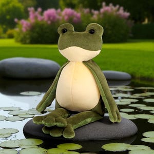 May include: A plush frog toy with a green body, cream-colored belly, and large black eyes. The frog is sitting on a gray stone in a pond with lily pads. The background features a blurred garden with pink flowers and green grass.