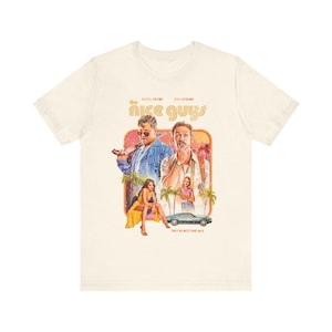 May include: Off-white t-shirt with a vintage-style movie poster design for "The Nice Guys." The graphic includes actors, a car, and palm trees, with the text "THE NICE GUYS" and "THEY'RE NOT THAT NICE."