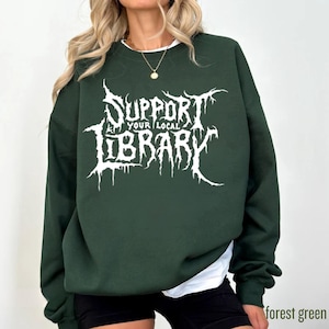 May include: A forest green sweatshirt featuring the white text "SUPPORT YOUR LOCAL LIBRARY". The text is in a stylized font. The sweatshirt has a crew neck and long sleeves. A white undershirt is also visible.