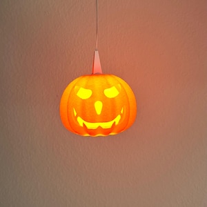 Könnte beinhalten: Eine orangefarbene Halloween-Kürbislaterne mit einem geschnitzten Gesicht hängt an einem dünnen weißen Kabel. Der Kürbis hat einen lächelnden Mund, dreieckige Augen und eine kleine Nase. Das Licht im Inneren erzeugt einen warmen Schein.