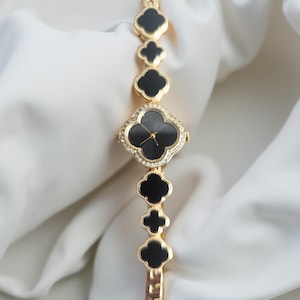 May include: Gold-coloured watch with a black clover-shaped face and a band featuring alternating black clover-shaped links. The watch face is surrounded by small, clear crystals. The background is a white, draped fabric.
