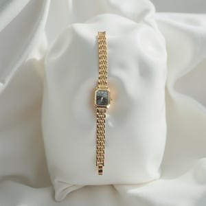 Peut inclure: Une montre-bracelet dorée avec un cadran noir rectangulaire et un bracelet à maillons. La montre est présentée sur une surface blanche texturée, mettant en valeur son design élégant. La montre comporte des chiffres romains.