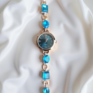 May include: A gold-toned wristwatch with a teal face and blue gemstone accents. The watch features a round face with gold hands and a quartz movement. The band is adorned with rectangular and oval-shaped blue gemstones.