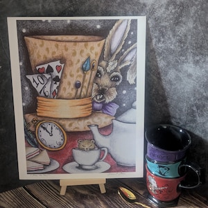 May include: An illustrated print with an Alice in Wonderland theme, showing a rabbit, playing cards, and a teacup with a mouse. A stack of colourful teacups and a golden spoon are also visible. The artwork is displayed on a small wooden easel.